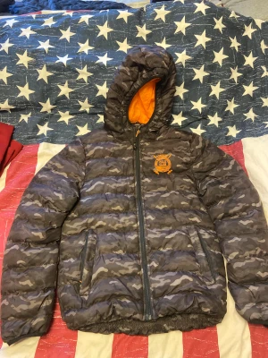 Camouflage pufferjacka från Vinson Polo Club - Säljer en mörkgrå pufferjacka med camouflagemönster från Vinson Polo Club. Jackan har huva med orange foder, broderad logga på bröstet och dragkedja framtill. Två sidofickor med dragkedja och elastiska muddar vid ärmslut. Perfekt för kalla dagar. Jackans zipper har krånglat sig lite men den funkar som den ska. Jackan överlag är i gott skick. Storlek S. 