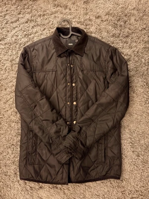 Quiltad brun svart jacka från ASOS - Quiltad brun svart jacka från ASOS med snygg manchesterkrage och tryckknappar framtill. Jackan har ett rutmönster, två sidofickor och långärmade ärmar. Perfekt för dig som gillar klassisk stil med modern touch.