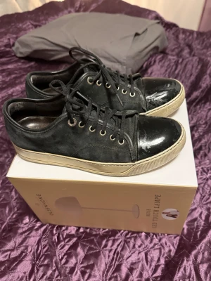 Svarta sneakers från Lanvin med lackad tå - Svarta sneakers från Lanvin med snygg lackad tå och snörning. Ovandelen är i mocka och skinn, medan sulan är beige och platt. Metallfärgade öljetter och rund tå ger en cool och stilren vibe. Perfekt för dig som gillar exklusiva detaljer och klassisk design. Har inte låda eller något annat