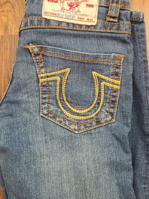 True Religion jeans med kontrastsömmar - Säljer ett par klassiska True Religion jeans i mörkblå denim med tydliga kontrastsömmar i beige. Modellen har raka ben och låg midja, samt ikoniska bakfickor med broderad logga. 73 cm innerbenlängd. 97 ytterbenlängd. 34 midjemått.  Skriv för frågor.