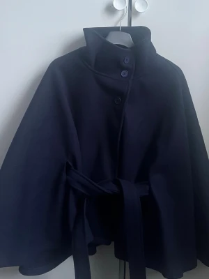 Mörkblå cape-coat med bälte - Säljer en mörkblå cape-kappa med hög krage, knappar framtill och matchande bälte i midjan. Jackan har en stilren och svepande siluett som ger en cool och elegant vibe. Perfekt för dig som vill sticka ut med en unik ytterjacka.