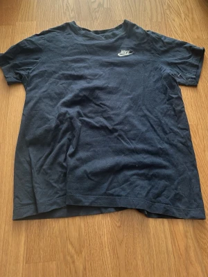 Svart Nike t-shirt i bomull - Klassisk svart t-shirt från Nike med broderad vit logga på bröstet. Tillverkad i mjuk bomull och har en normal passform med korta ärmar. Perfekt till chill vardagsstil eller när du vill köra på en enkel look.