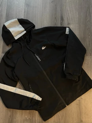 Svart vindjacka från Lacoste - Säljer en svart vindjacka från Lacoste med vita reflexdetaljer på huva, ärmar och rygg. Jackan har dragkedja framtill, två fickor med dragkedja och justerbar huva. Ikoniska Lacoste-loggan på bröstet och cool sportig look. Storleken på jackan är M i junior 