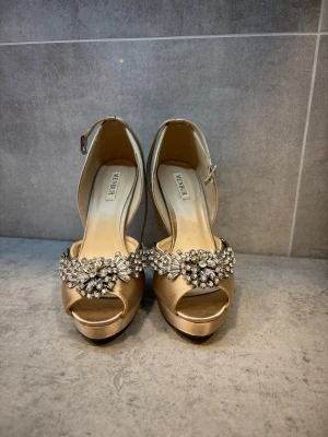Guldiga pumps med strass från Menbur - Snygga pumps i guldigt satin från Menbur med öppen tå och hög klack. Skorna har en bred rem med glittrande strassdekor framtill och justerbar rem runt ankeln. Perfekta för dig som vill sticka ut med elegans och bling.