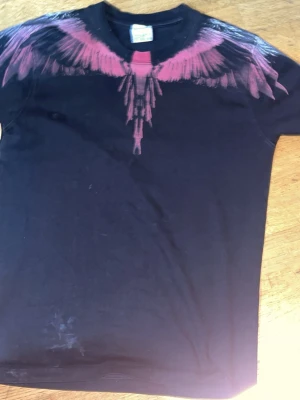 Svart t-shirt med röd sträckor från Marcelo burlon  - Marcelo burlon Säljer en svart t-shirt med ett fett röd tryck som liknar fjädrar eller vingar över axlarna och ner mot bröstet. T-shirten har rund hals och korta ärmar. Materialet känns mjukt och skönt, troligtvis bomull.