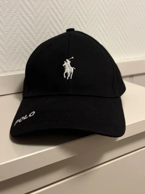 Svart Polo Ralph Lauren keps - Snygg svart keps från Polo Ralph Lauren med klassisk broderad logga i vitt framtill och justerbart spänne bak. Kepsen har böjd skärm och Polo-tryck på insidan. Perfekt accessoar för en chill och sportig look.