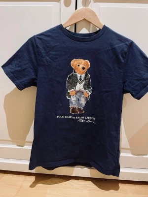 Mörkblå Polo Bear t-shirt Ralph Lauren - Säljer en mörkblå t-shirt från Polo Ralph Lauren med ikoniskt Polo Bear-tryck på bröstet. T-shirten har rund hals, korta ärmar och är gjord i mjuk bomull. Perfekt för dig som gillar klassisk streetstyle med en twist.