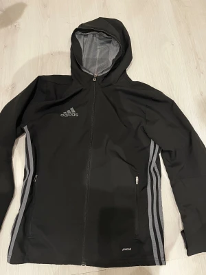 Vindjacka  - Svart vindjacka från Adidas i storlek XS med huva och hel dragkedja. Jackan har grå ränder längs sidorna, två fickor med dragkedja och meshfoder. Perfekt för träning och aktiva dagar, med ventilerande detaljer på ryggen och snygg sportig look.
