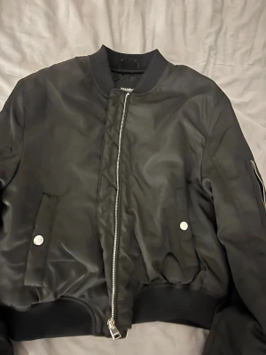 Svart bomberjacka från Pull&Bear - Säljer en svart bomberjacka från Pull&Bear med klassisk ribbad krage och muddar. Jackan har dragkedja framtill, fickor med tryckknappar och en extra ficka med dragkedja på ärmen. Materialet är glansigt och känns lätt, perfekt för en cool streetstil. 