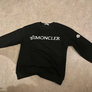 Svart Moncler sweatshirt med logga - Svart sweatshirt från Moncler med stor vit logga framtill och klassisk Moncler-patch på ärmen. Tröjan har rund halsringning och långa ärmar, perfekt för en clean och stilren look. Tillverkad i mjuk bomull för maximal komfort. Perfekt till sommaren och till hösten/ vintern. Passar dig som har storlek s eller m