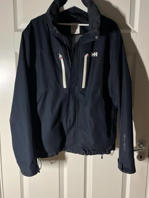 Helly Hansen vindjacka - Säljer en marinblå vindjacka från Helly Hansen i storlek L men passar även M användare. Jackan har två vita dragkedjor framtill, hög krage och justerbar nederkant. Materialet är slitstark polyester. Vid fler frågor hör av er!
