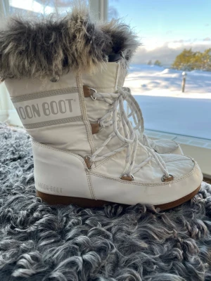 Vita Moon Boots med fuskpäls - Säljer ett par vita Ikoniska Moon Boots med snörning och brun sula. Skorna har en fluffig kant i fuskpäls upptill och är perfekta för kalla vinterdagar. Tillverkade i syntetmaterial och designade för att hålla värmen. Innersula går att knäppa upp och ta ur för att vid behov t.ex torka. Klassisk Moon Boot-logga på sidan. Nylonkant på högerstövelns överdel är något avskavd men det har ingen påverkan på funktionen. 