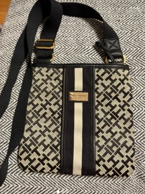 Mönstrad axelväska Tommy Hilfiger - Snygg axelväska från Tommy Hilfiger med svart och beige geometriskt mönster och bred axelrem. Väskan har en dekorativ rand i mitten och guldfärgad logga framtill. Perfekt storlek för det viktigaste och enkel att bära crossbody.