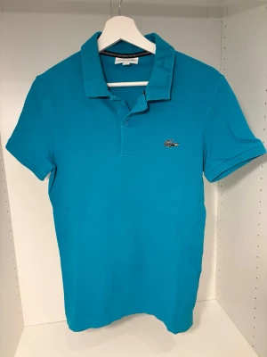 Turkos pikétröja Lacoste slim fit XS - Säljer en turkos pikétröja från Lacoste i slim fit-modell, storlek XS. Klassisk krage med två knappar och den ikoniska krokodilloggan på bröstet. Tröjan är kortärmad och tillverkad i bomull för en skön känsla.