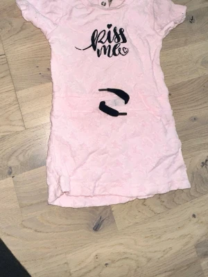 Ljusrosa topp Kiss Me från Dear Body D-XEL rosa klänning, storlek 74/80 - Supersöt ljusrosa topp från Dear Body med svart text 'Kiss Me' framtill. Kortärmad modell i mjukt bomullstyg med diskret struktur. Perfekt för dig som gillar gulliga och lekfulla plagg. Rund halsringning och normal passform.Söt rosa klänning med korta ärmar och texten "Kiss me".