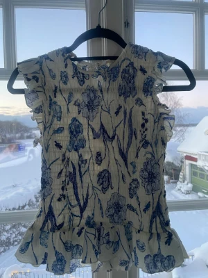 Blommig blus med volang från H&M - Säljer en söt blommig blus från H&M i storlek S. Blusen är vit med blått blommönster, har volangdetaljer vid ärmar och nederkant samt är ärmlös. Materialet är lätt och krinklat, perfekt för vår och sommar. Nyckelhålsöppning bak i nacken.