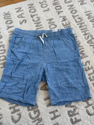 Blå linneshorts med snörning - Säljer ett par blå linneshorts från H&M med elastisk midja och vit snörning framtill. Shortsen har sidofickor och en bakficka, perfekt för chill dagar. Materialet är svalt och passar grymt till sommaren.