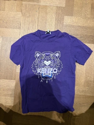 Lila Kenzo t-shirt med tigertryck - Kenzo t-shirt i lila med ikoniskt tigertryck och texten 'Kenzo Paris' på bröstet. Klassisk rund hals och korta ärmar. Tillverkad i mjuk bomull som känns skön mot huden. Perfekt för dig som vill sticka ut med en cool streetwear vibe.