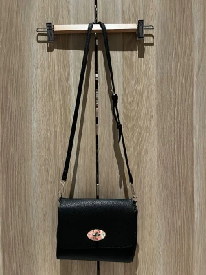 Svart axelväska med justerbart band - Säljer en stilren svart axelväska i syntet med justerbart axelband och silverfärgade detaljer. Väskan har ett klassiskt lock med vridlås framtill och en praktisk dragkedjeficka på baksidan. Insidan har flera fack för bra organisering.