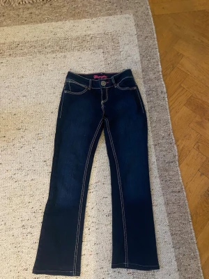 Mörkblå Wrangler jeans med broderi - Snygga mörkblå jeans från Wrangler med kontrastsömmar och broderade bakfickor. Klassisk femficksmodell med raka ben och låg midja. Tuffa detaljer med synlig logga och dekorativa sömmar. Perfekta för dig som gillar en tidlös jeanslook. Skulle säga xs i midjan med dom byxorn är till en kort person innerbenslängden är 69 och midja 33 bra för priset 