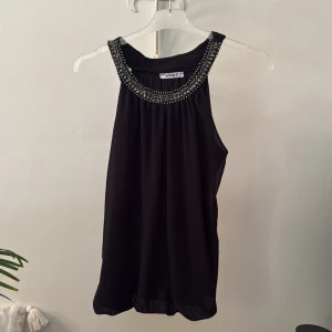 Svart ärmlös topp med pärldetaljer - Svart ärmlös topp från MYHOLLS i luftig polyester. Halsringningen är dekorerad med glittriga pärlor och stenar som ger en lyxig känsla. Modellen är lös och faller snyggt.