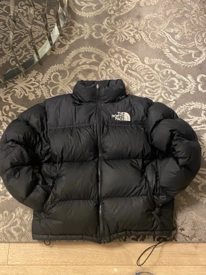 Svart pufferjacka från The North Face - Säljer en svart pufferjacka från The North Face i storlek M. Jackan har hög krage, dragkedja framtill och två fickor med dragkedja. Klassisk logga på bröstet och ryggen. Perfekt för kalla dagar och riktigt skön att bära. Ny pris runt 3000. Finns kvitto. 