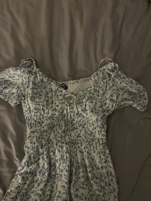 Blommig kortklänning från Bik Bok - Supersöt kortklänning från Bik Bok i vit med blått blommönster. Klänningen har puffiga kortärmar, smock över byst och midja samt en söt knytning vid urringningen. Perfekt för sommarens alla dagar! Har använt 1 gång!