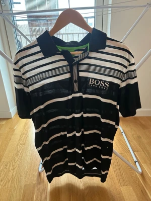 Randig pikétröja från Hugo Boss - Snygg svart och vit randig pikétröja från Hugo Boss med klassisk krage och knappslå. Tröjan har broderad logga på bröstet och är tillverkad i ett lätt och bekvämt Nanotex-material. Perfekt för en stilren och sportig look.
