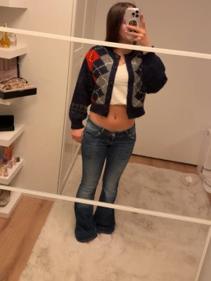 Mörkblå croppad kofta med rutor - Säljer en croppad kofta i mörkblått med rutigt argylemönster i rött, grått och vitt. Koftan har långa ärmar, knappar framtill och är stickad i ett mjukt material. Perfekt för lager-på-lager och snygg till jeans.