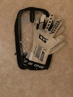 Vita målvaktshandskar från The One - Säljer ett par vita målvaktshandskar från The One med svart logga och detaljer. Handskarna har elastisk mudd med texten 'ONE' och greppvänlig insida. Perfekta för dig som vill ta ditt spel till nästa nivå på fotbollsplanen.