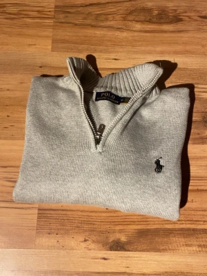 Grå stickad tröja Polo Ralph Lauren - Snygg grå stickad tröja från Polo Ralph Lauren med halv dragkedja och ribbad krage. Klassisk logga broderad på bröstet. Perfekt till lager-på-lager och passar både till jeans och chinos. Långärmad och i mjukt material.