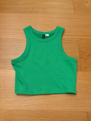 Grön ribbad linnetopp från H&M Divided, storlek S - Snygg grön croppad linnetopp från H&M Divided i storlek S i ribbad bomull. Toppen är ärmlös med rund halsringning och har en tight passform som passar perfekt till sommaren. Enkel och stilren design som är lätt att matcha. 94% Polyester, 6% Elastan.
