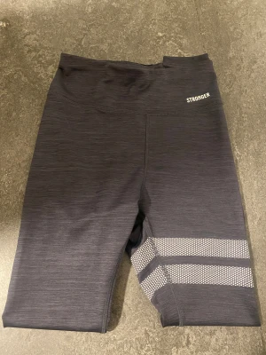 Svarta leggings från Stronger  - Säljer ett par svarta leggings från Stronger i storlek XS. De har vita prickiga ränder vid benen och är tillverkade i stretchigt material med 73% polyester och 22% elastan. Perfekta för träning och riktigt sköna att ha på sig.