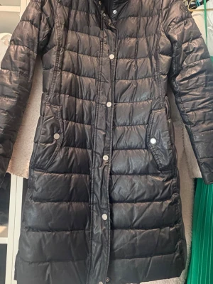 Svart lång pufferjacka Tommy Hilfiger - Säljer en svart lång pufferjacka från Tommy Hilfiger med quiltad design och tryckknappar framtill. Jackan har två fickor med knappstängning och broderad logga på ärmen. Perfekt för kalla dagar och riktigt snygg till streetwear.