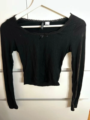 Svart lace cami med rosett - Säljer en svart ribbad långärmad topp från H&M Divided i storlek XS. Toppen har spetskant vid ringningen och en liten rosett framtill. Materialet är mjukt, stretchigt och skönt på kroppen.