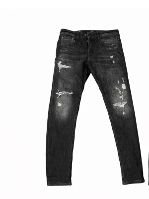 Replay svarta  jeans (willbi) - Säljer ett par svarta Replay jeans med slitna detaljer och trasiga partier framtill. Jeansen har fem fickor, knappgylf och en smal passform. Materialet är klassisk jeans i bomull och de har en cool, mörk tvätt med slitningar för en edgy look.