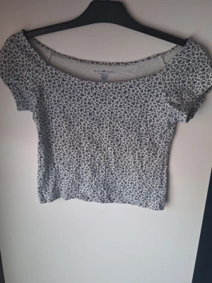 Blommig topp från Brandy Melville - Supersöt topp från Brandy Melville med blått blommönster på vit botten. Modellen är croppad med korta ärmar och off shoulder halsringning. 