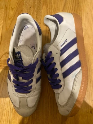 Adidas Samba vita och lila sneakers - Säljer ett par klassiska Adidas Samba sneakers i vitt skinn med lila detaljer och snörning. Skorna har de ikoniska tre ränderna i lila på sidorna och en brun gummisula. Perfekta för dig som gillar retrostil och streetwear.