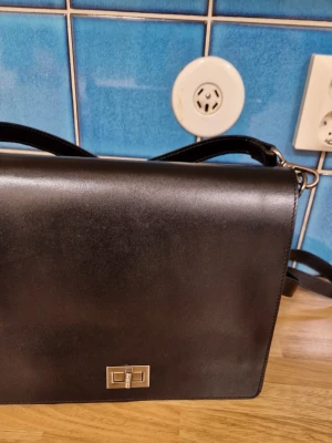 Svart axelremsväska i skinn - Stilren svart axelremsväska i shiny skinn med silverfärgat spänne framtill. Väskan har justerbar rem, flera fack och dragkedja på baksidan. Perfekt för dig som vill ha en klassisk och praktisk väska med snygg finish.