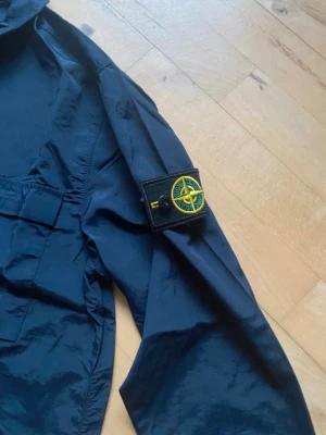 Svart vindjacka från Stone Island - Säljer en svart vindjacka från Stone Island med klassisk patch på ärmen. Jackan har dragkedja framtill, två stora bröstfickor och en enkel krage. Materialet är lätt och känns syntetiskt, perfekt för blåsiga dagar. Stilen är normal passform och jackan har långa ärmar.