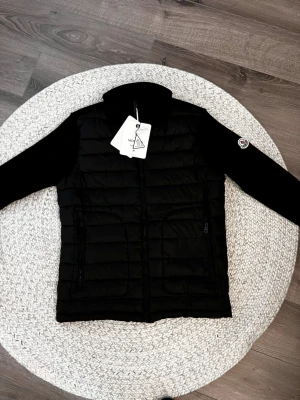 Moncler cardigan svart - Snygg svart cardigan från Moncler i storlek M. Jackan har quiltad kropp i syntetmaterial och stickade ärmar med Moncler-logga på armen. Hög krage och dragkedja framtill. Perfekt för dig som vill ha en clean och modern look.