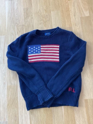 Mörkblå stickad tröja Polo Ralph Lauren - Mörkblå stickad tröja från Polo Ralph Lauren med amerikansk flagga i rött, vitt och blått på bröstet. RL-broderi i rött vid nederkanten. Klassisk rund hals och långa ärmar. 