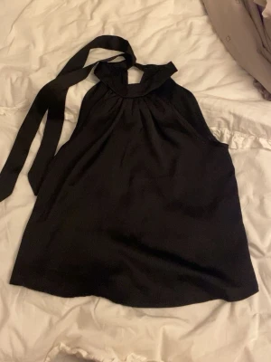 Svart halterneck linnetopp med knyt - Säljer en svart halterneck linnetopp med breda band som knyts i nacken. Toppen har en lös passform och är tillverkad i ett mjukt, glansigt material som ger en elegant känsla. Perfekt till sommarens alla tillfällen!