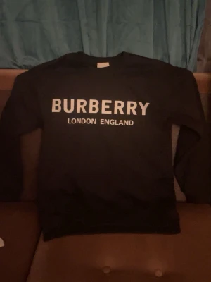 Svart sweatshirt från Burberry - Säljer en svart sweatshirt från Burberry med stor vit logga och texten 'London England' på bröstet. Tröjan har rund hals och långa ärmar, perfekt för dig som vill ha en clean och stilren look. Materialet känns mjukt och skönt mot huden, säljer den då den inte passar mig. PRISET ÄR INTE HUGGET I STEN