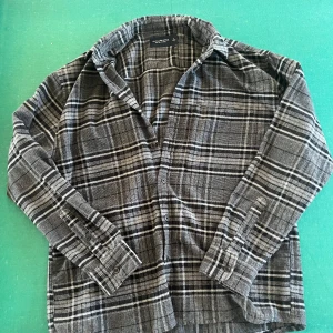 Rutig overshirt från Abercrombie & Fitch - Säljer en mjuk och skön overshirt från Abercrombie & Fitch i svart, grått och vitt rutmönster. Flanellmaterialet gör den extra bekväm och perfekt att slänga på sig över en t-shirt. Klassisk krage, knäppning framtill och långa ärmar.