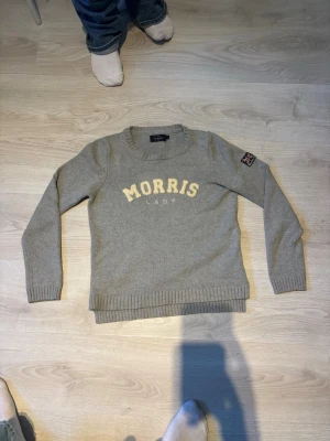 Grå stickad tröja från Morris Lady - Säljer en grå stickad tröja från Morris Lady med krämfärgad logga framtill och en liten patch på ärmen. Tröjan har rund hals, ribbade muddar och lång ärm. Perfekt till jeans för en chill och snygg look.