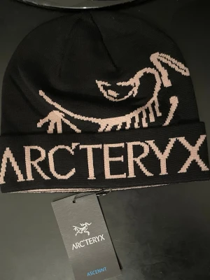Svart Arc'teryx mössa, Aldrig Använt! - Snygg svart mössa från Arc'teryx med stor, stickad logga och fossilmotiv i ljusbeige. Mössan är tillverkad i mjukt stickat material och har en bred uppvikt kant. Perfekt för kalla dagar och outdoor-aktiviteter.