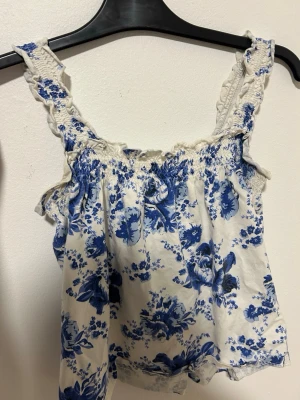 Blommig linnetopp från Kappahl XS - Supersöt linnetopp från Kappahl i vitt med blått blommönster. Toppen har breda axelband med volangdetaljer och smock upptill för en bekväm passform. Perfekt för varma dagar och ger en fräsch sommarkänsla.