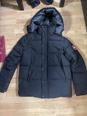 Svart pufferjacka från Canada Goose - Säljer en svart pufferjacka från Canada Goose med stor huva och klassisk logga på ärmen. Jackan har dragkedja och tryckknappar framtill samt två snedställda fickor. Perfekt för kalla dagar och riktigt snygg till vinterlooken.