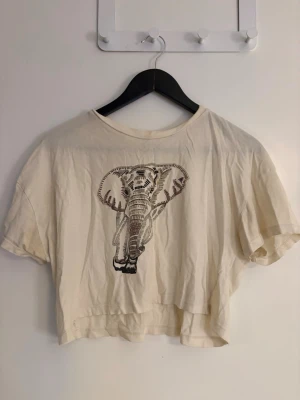 Dam krämvit croppad t-shirt med elefant - Säljer en krämvit croppad t-shirt med ett stort tryck av en elefant på framsidan. T-shirten har korta ärmar i storlek XS. Perfekt för dig som gillar djurmotiv och vill ha en chill vibe på outfiten.